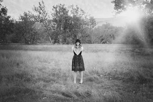 Black and white image of a woman in a plaid skirt standing alone in a misty meadow with soft sunbeams and trees behind her.