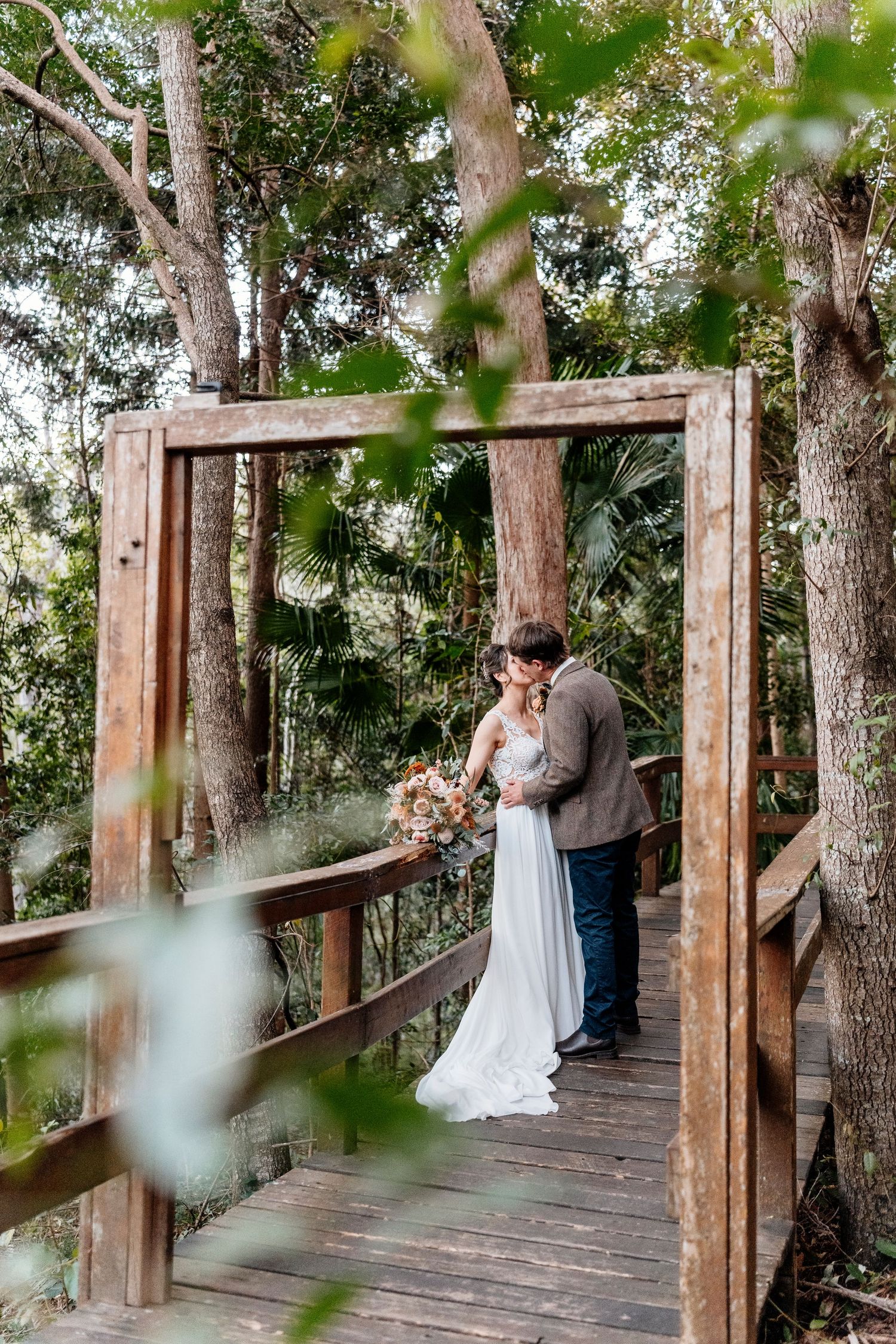 Bride and groom share a romantic kiss on a wooden bridge surrounded by lush forest trees during their outdoor wedding at walkabout creek