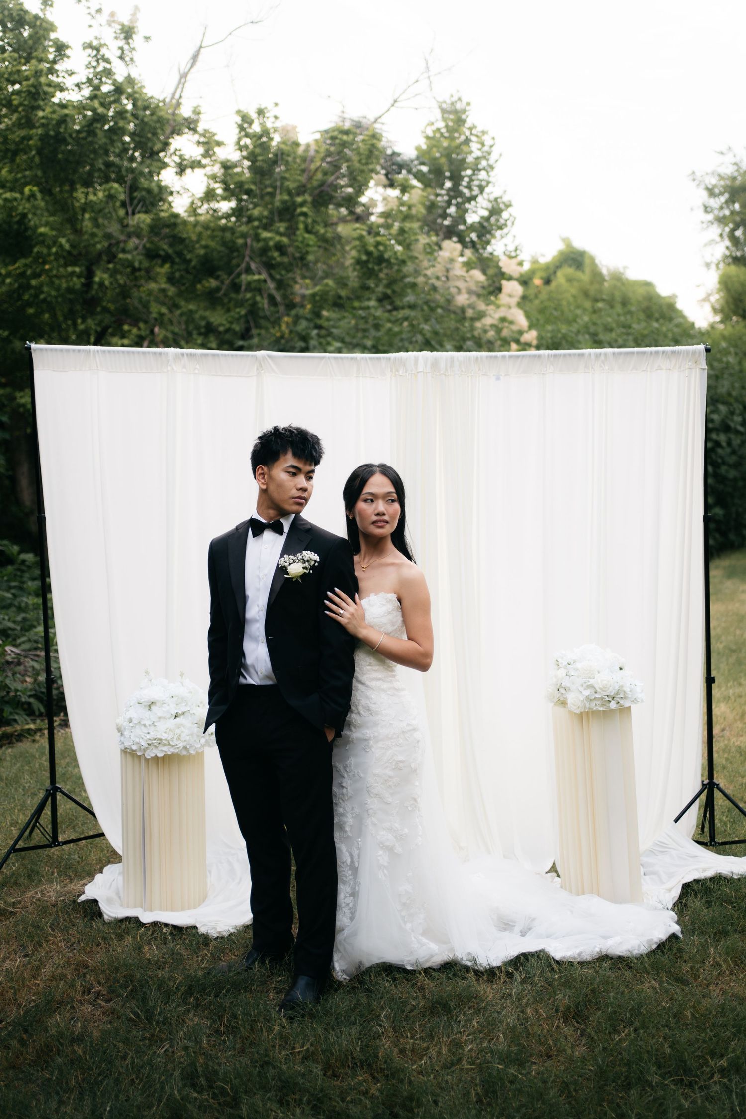 Bride and groom pose together in front of a white draped backdrop with floral pedestals at outdoor wedding.