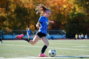 Soccer players chase ball on turf field with fall foliage in background.