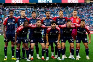 Soccer team poses for traditional pre-match lineup photo wearing dark uniforms on stadium field.