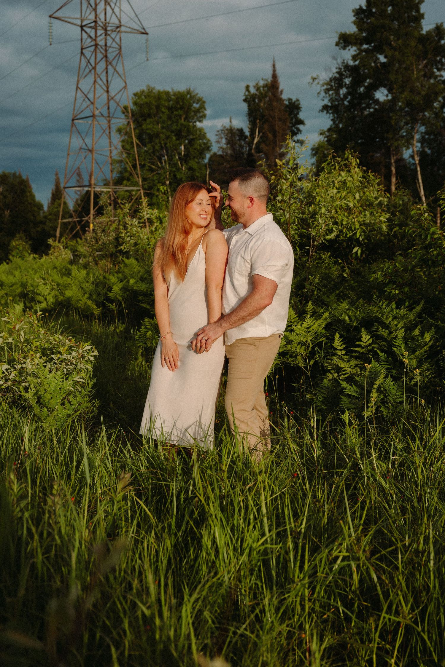 Couple holds hands in lush green field with power tower and dramatic cloudy sky at golden hour.