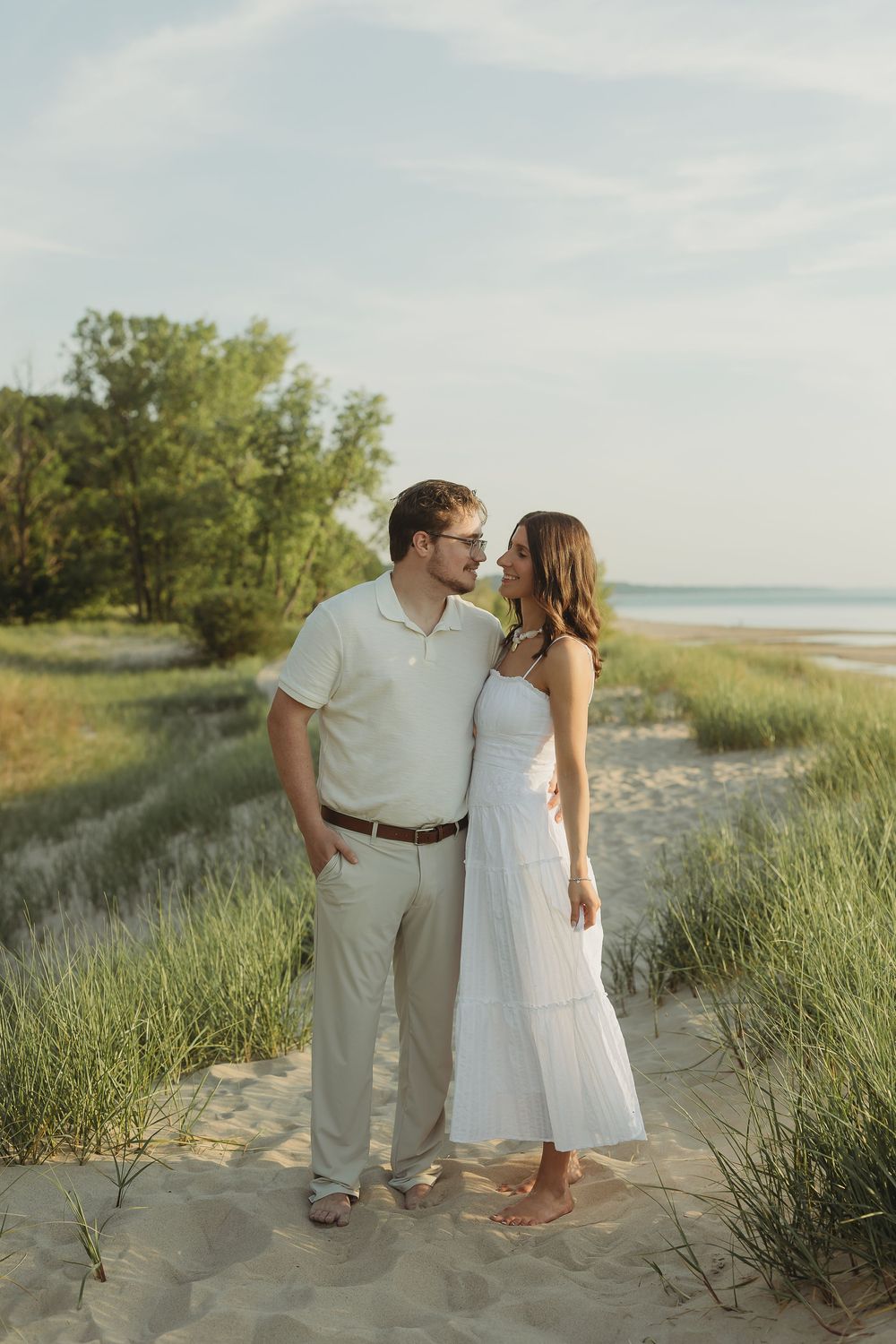 Couple in white outfits sharing a romantic moment on a sandy beach path with green dunes and ocean in the background.