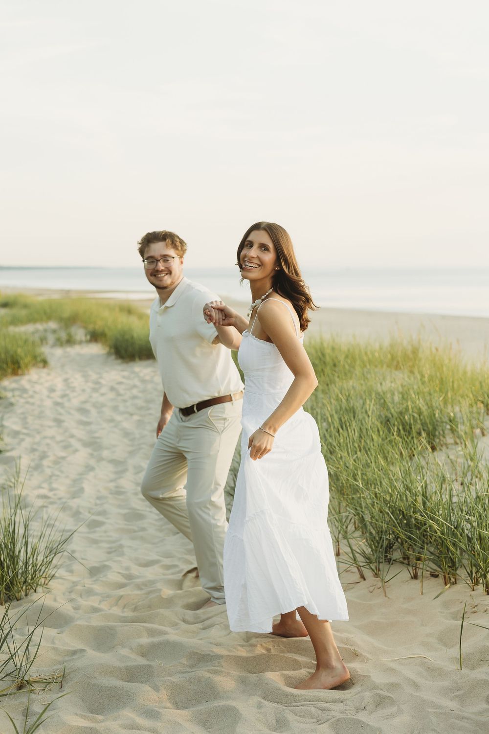 A smiling couple walks hand in hand on a sandy beach, dressed in white and beige outfits with ocean grass nearby.