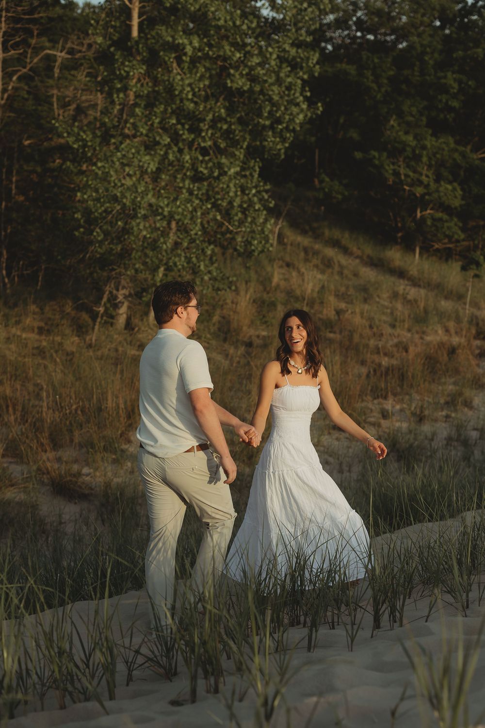 Couple holding hands walking on sandy dunes at golden hour, woman in white dress, man in casual attire, trees in background.