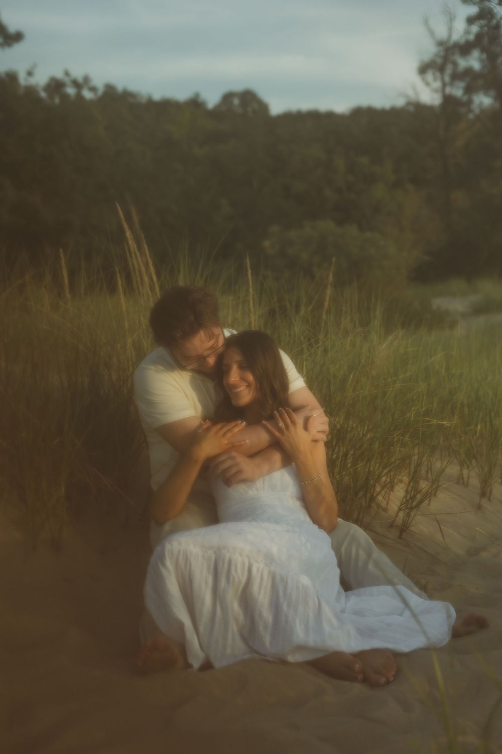 A couple embraces romantically on sandy dunes, woman in white dress, surrounded by golden grass and trees at sunset.