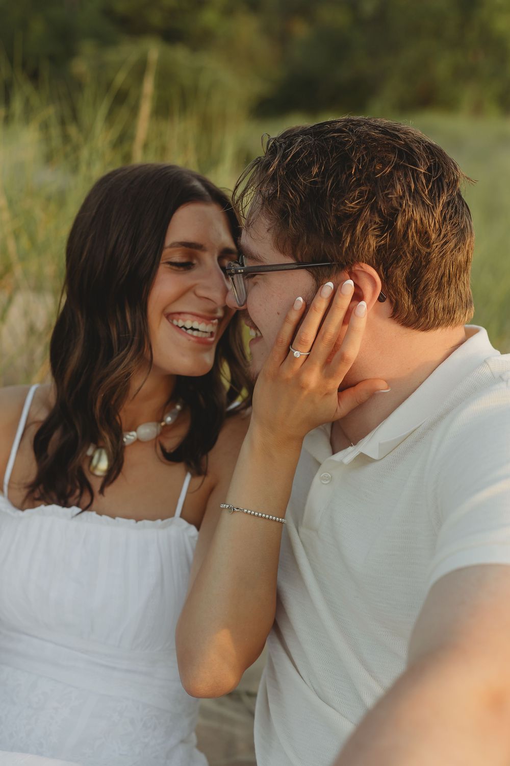 A smiling couple shares a tender moment outdoors, woman in white dress touching mans face, wearing matching bracelets.