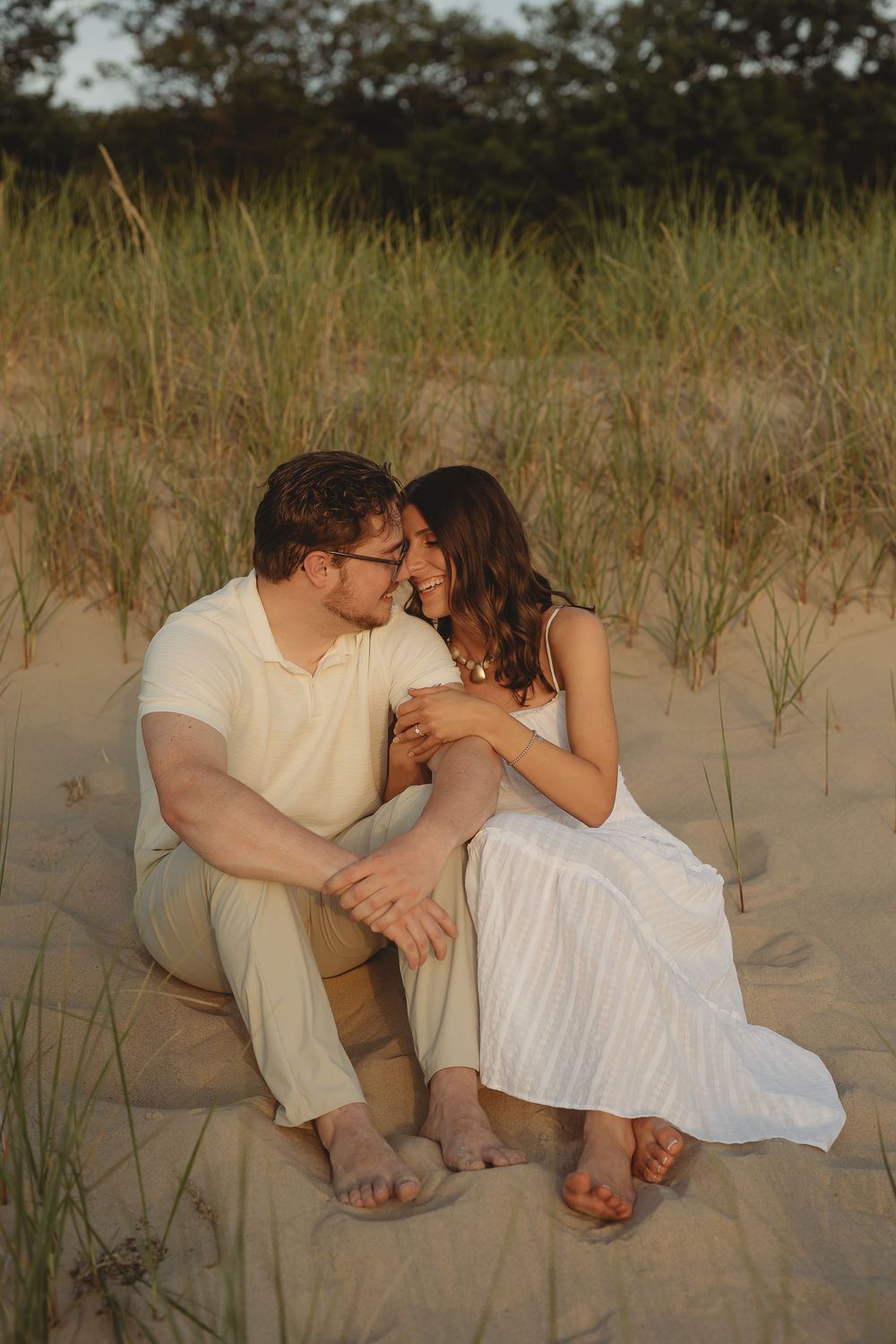 Couple sitting together on sandy beach dunes at golden hour, sharing an intimate moment in casual white and tan outfits.