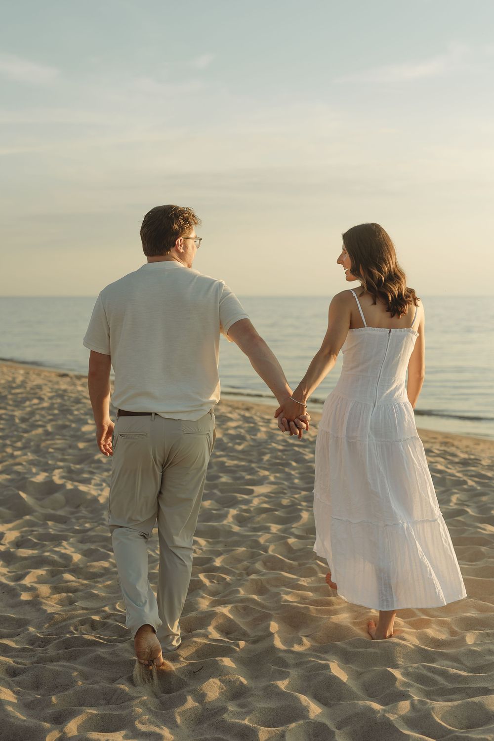 Couple holding hands walking on sandy beach at sunset, woman in white dress and man in casual attire, viewed from behind.