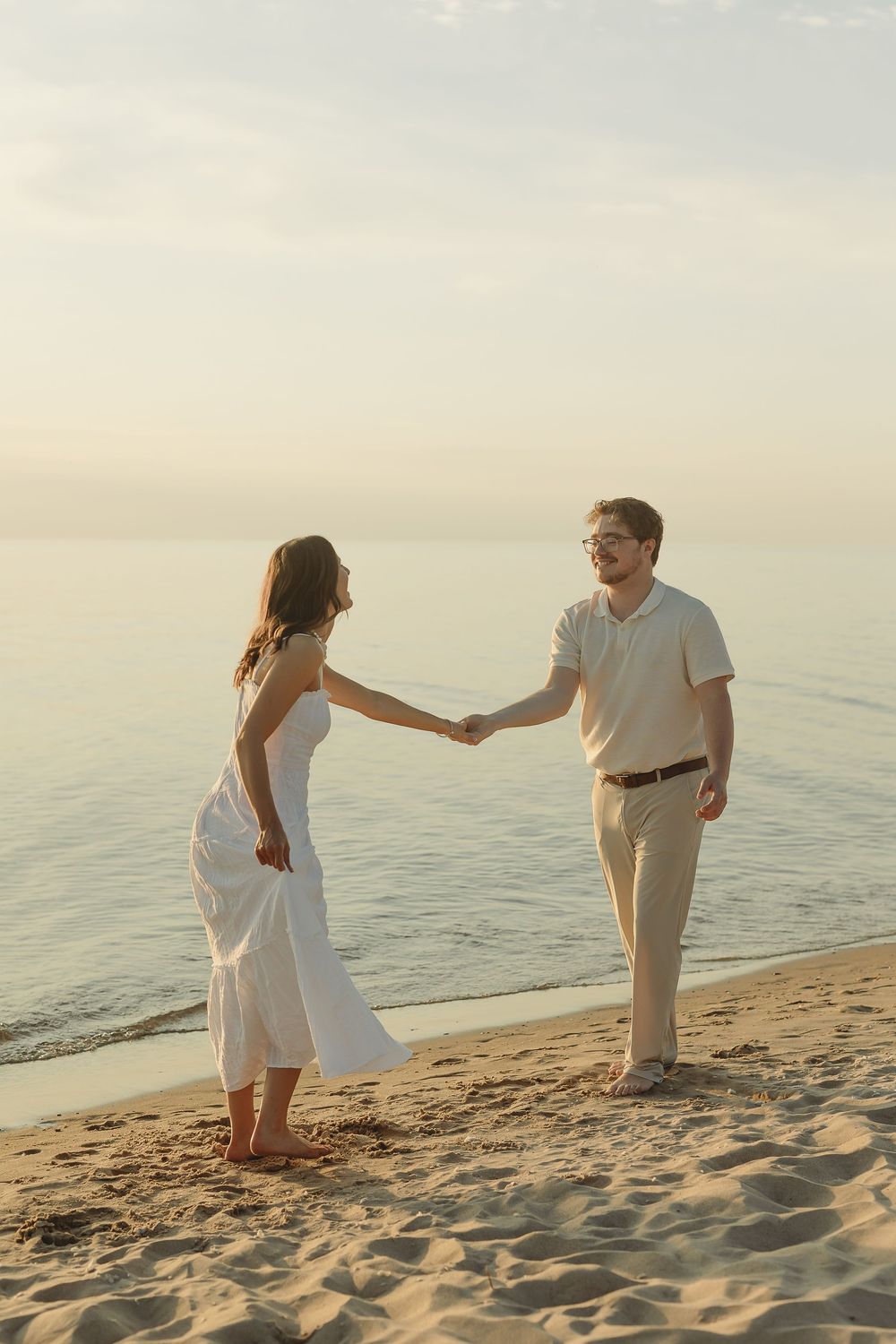 Couple holding hands on sandy beach at sunset, woman in white dress and man in light outfit facing each other.