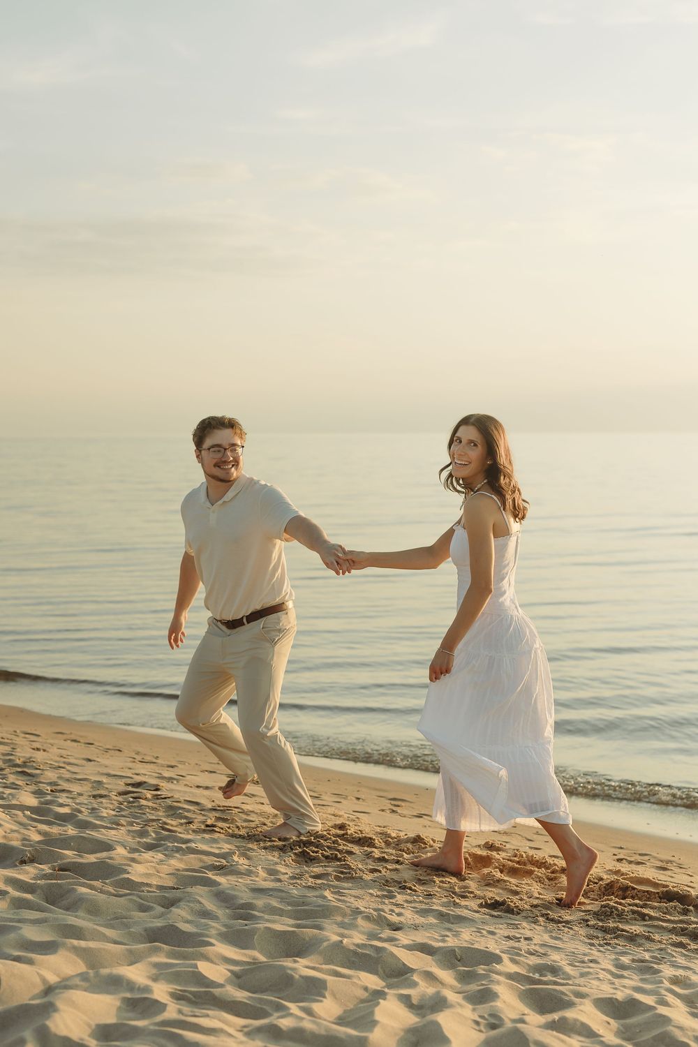 Happy couple holding hands and walking barefoot along a sandy beach at sunset in white outfits.