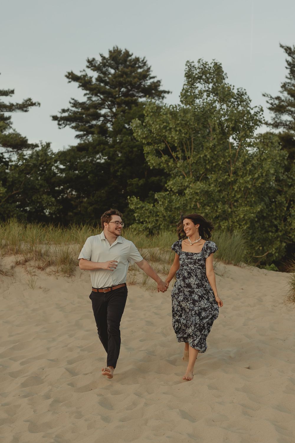 A couple holds hands and walks barefoot on sandy dunes, surrounded by lush green trees, smiling joyfully.