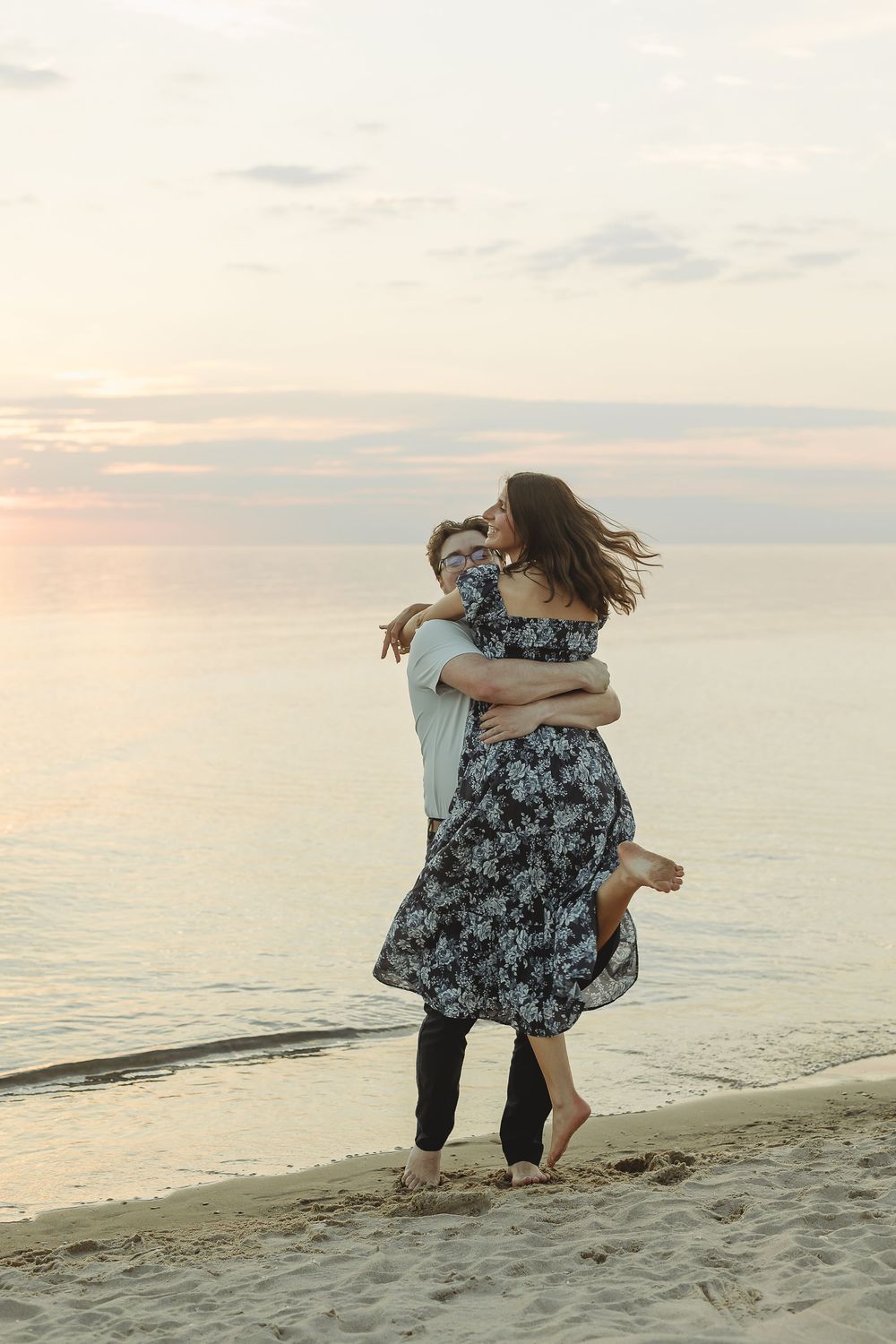 Couple embracing on a sandy beach at sunset, woman in floral dress lifted off ground near calm ocean waters.