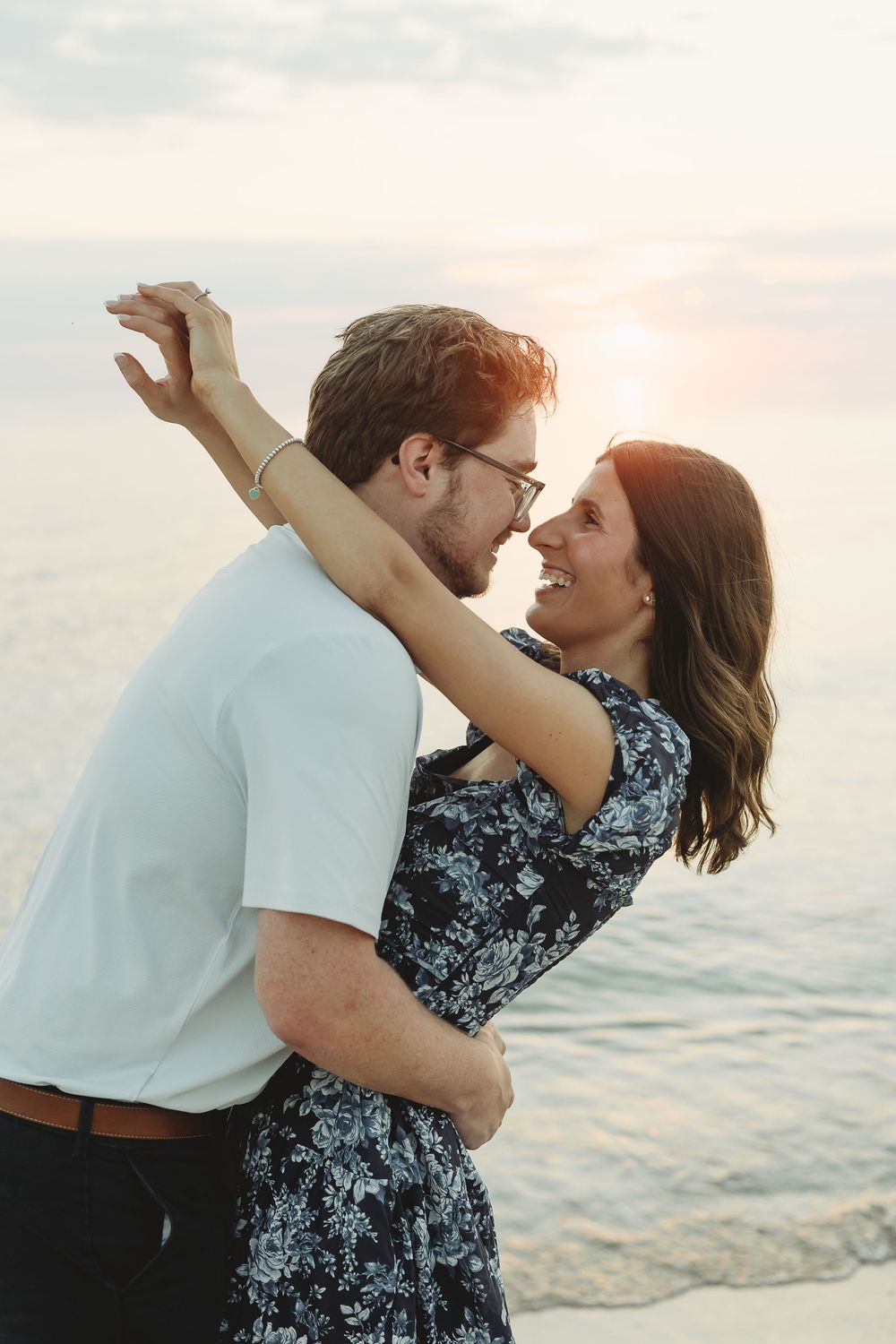 Couple embracing and smiling at each other on a beach at sunset, woman in floral dress with arms around mans neck.