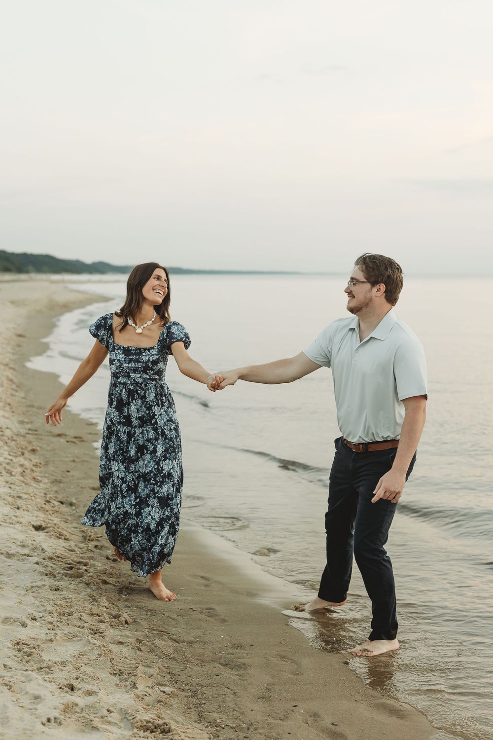 A couple holds hands and walks barefoot along a sandy beach at dusk, smiling at each other in casual summer outfits.