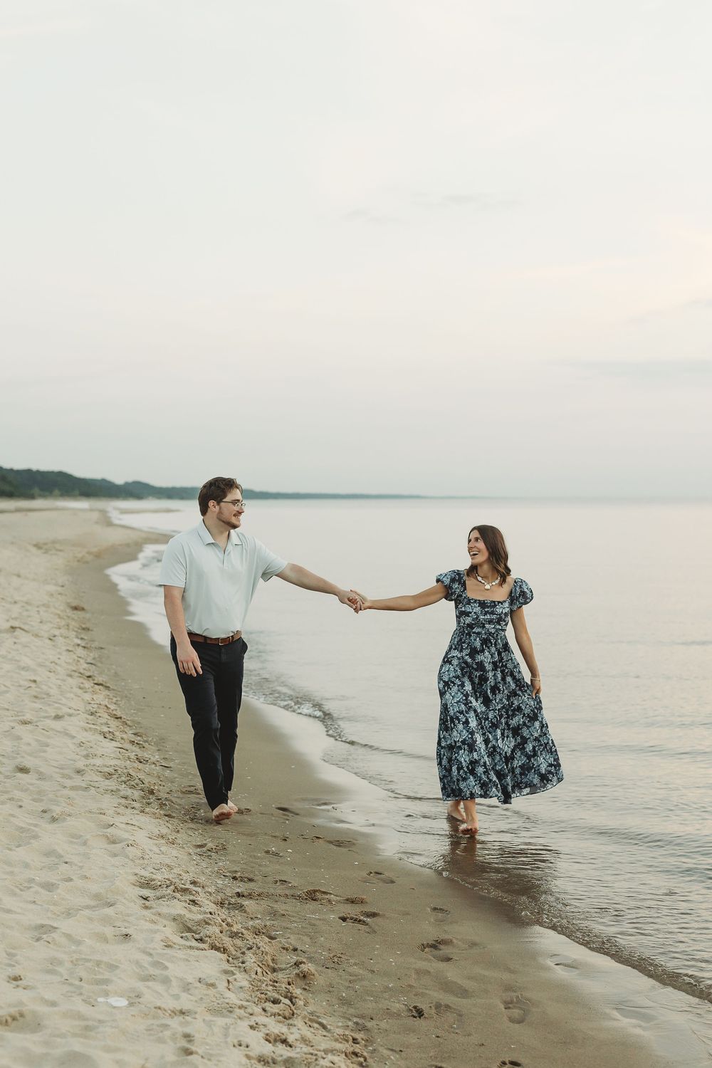 Couple holding hands walking barefoot along sandy beach shoreline at sunset in romantic outdoor engagement photo.