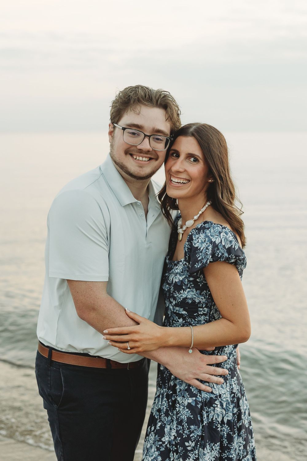 A smiling couple embraces by the ocean, man in light polo and glasses, woman in floral blue dress with pearl necklace.