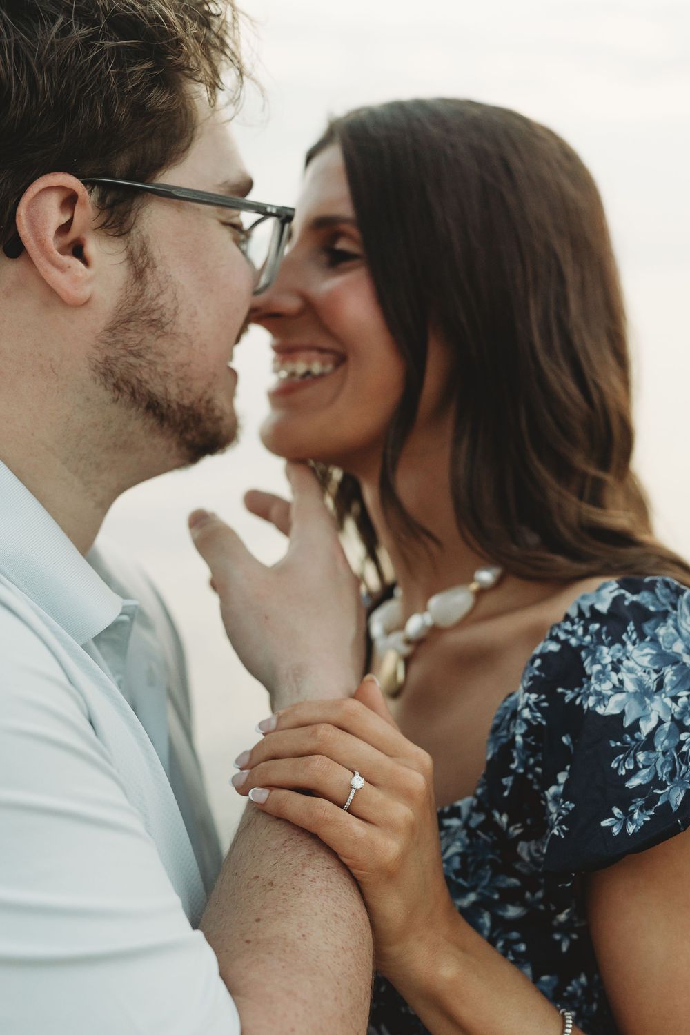 Smiling couple in romantic moment, woman in floral dress with engagement ring, man in glasses, soft outdoor lighting.