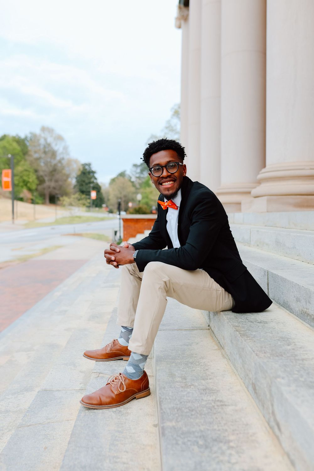 Stylish young man in black blazer and orange bow tie sits smiling on stone steps outside a building.