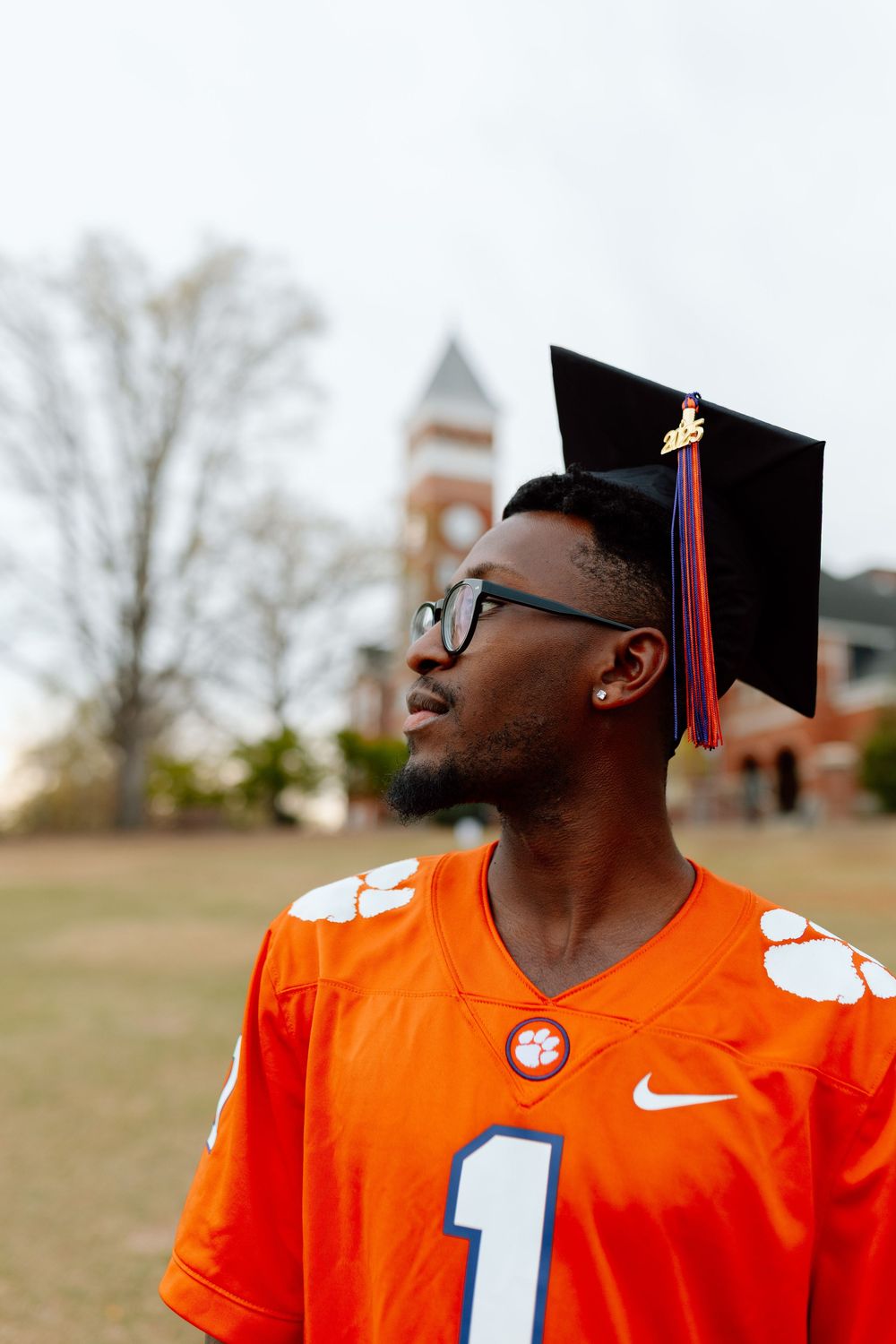 Graduate wearing Clemson orange jersey and black cap stands outdoors with campus clock tower in background.