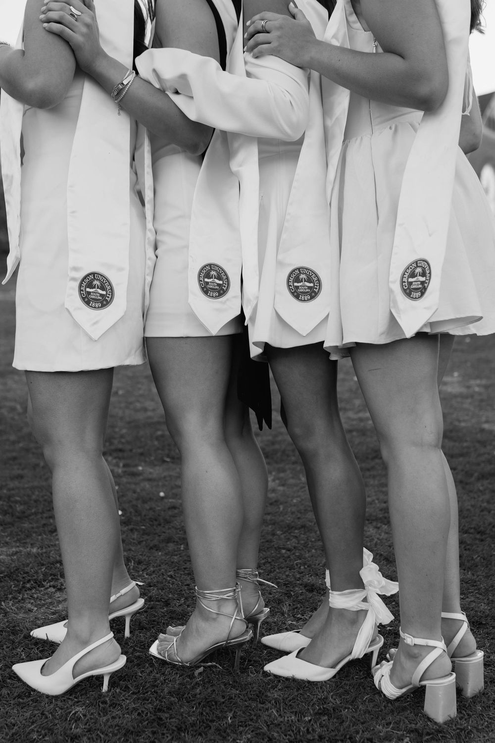 Black and white photo of graduates in white dresses and stoles showing university seals and heels.