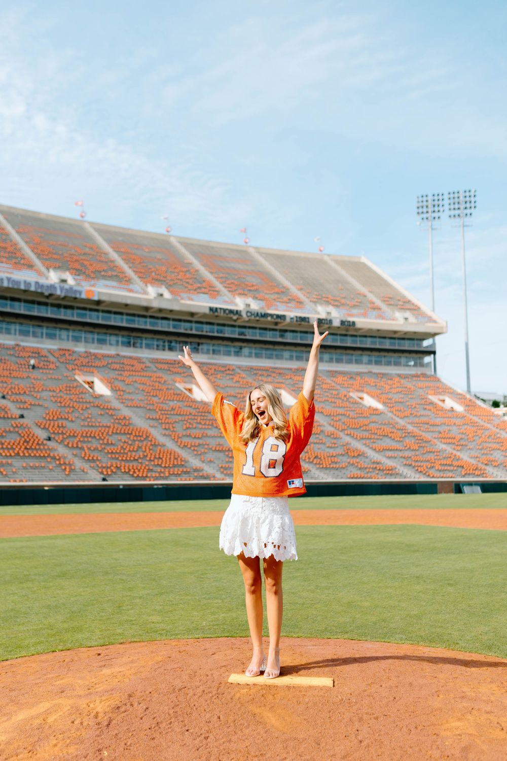 Young woman in orange jersey and white skirt celebrates on baseball pitcher's mound at Clemson University stadium.