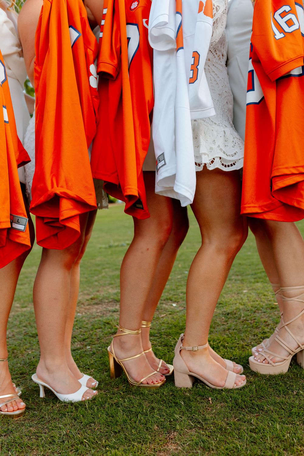 Group of women holding orange Clemson football jerseys wearing heels and white dresses on grass.