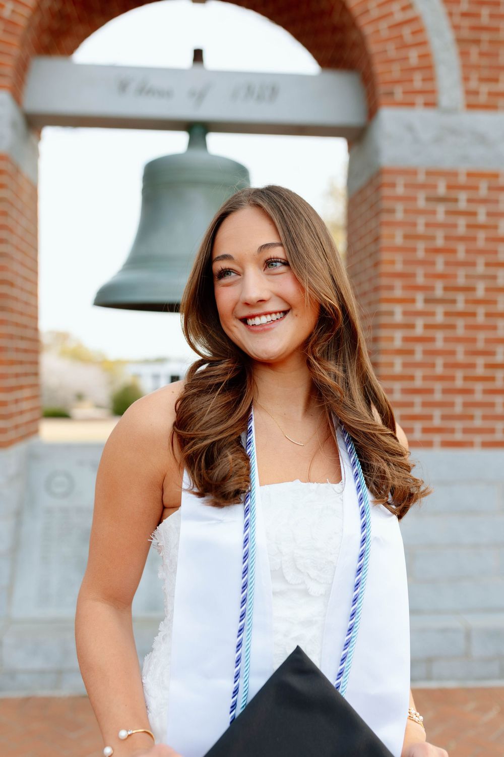 Smiling female graduate in white dress holds cap near campus bell tower arch at university.