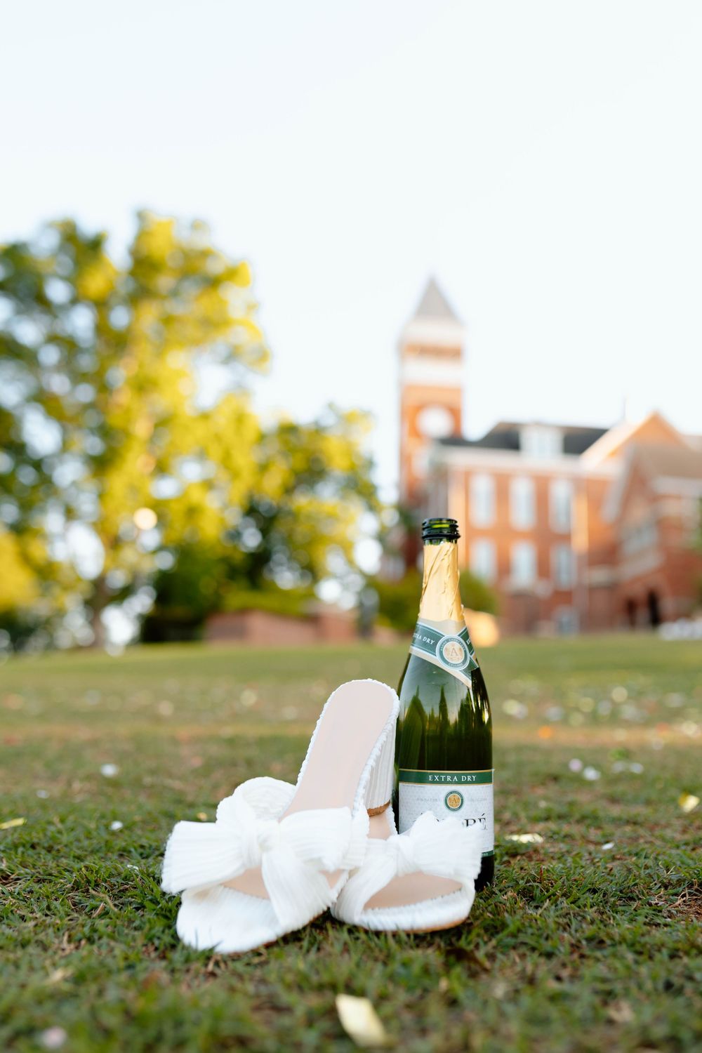 White heels and champagne bottle on grass with university clock tower in background.