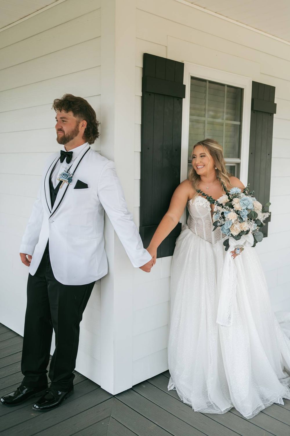 Bride in white tulle gown holding bouquet and groom in white tuxedo jacket hold hands outside a white building.
