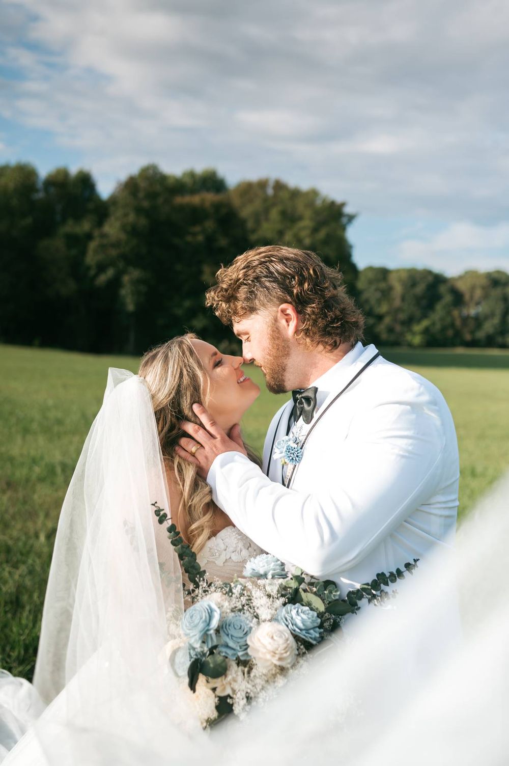 A bride and groom share a romantic kiss in a green field, bride holding blue and white bouquet, groom in white suit.