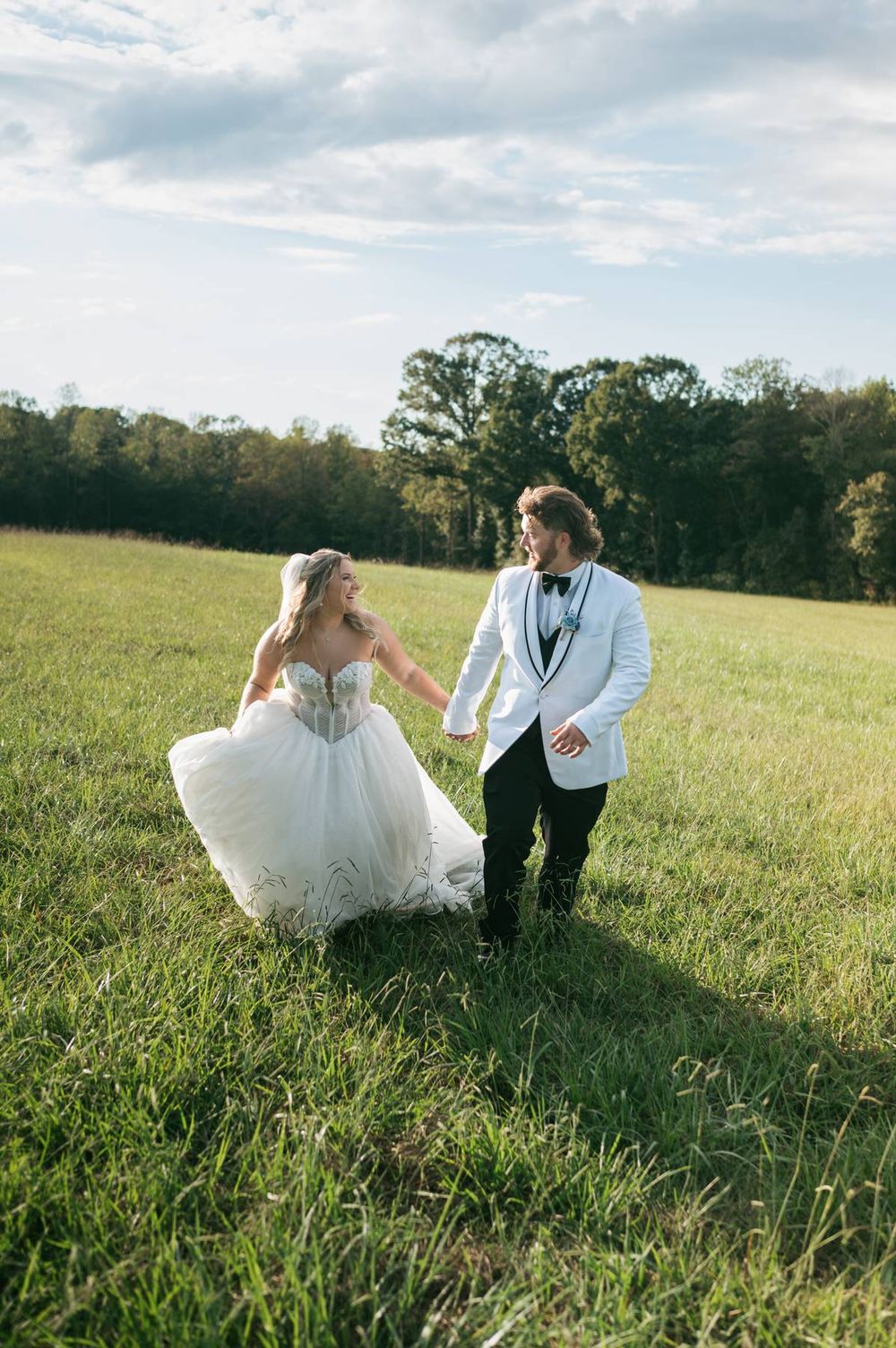 Bride in white ball gown and groom in white tuxedo hold hands walking through a lush green field on their wedding day.