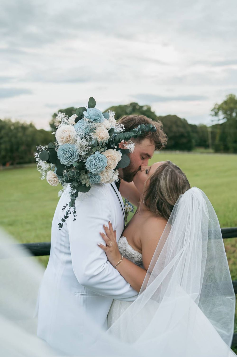 Bride and groom share a kiss holding a blue and white floral bouquet, with a flowing veil and green landscape backdrop.