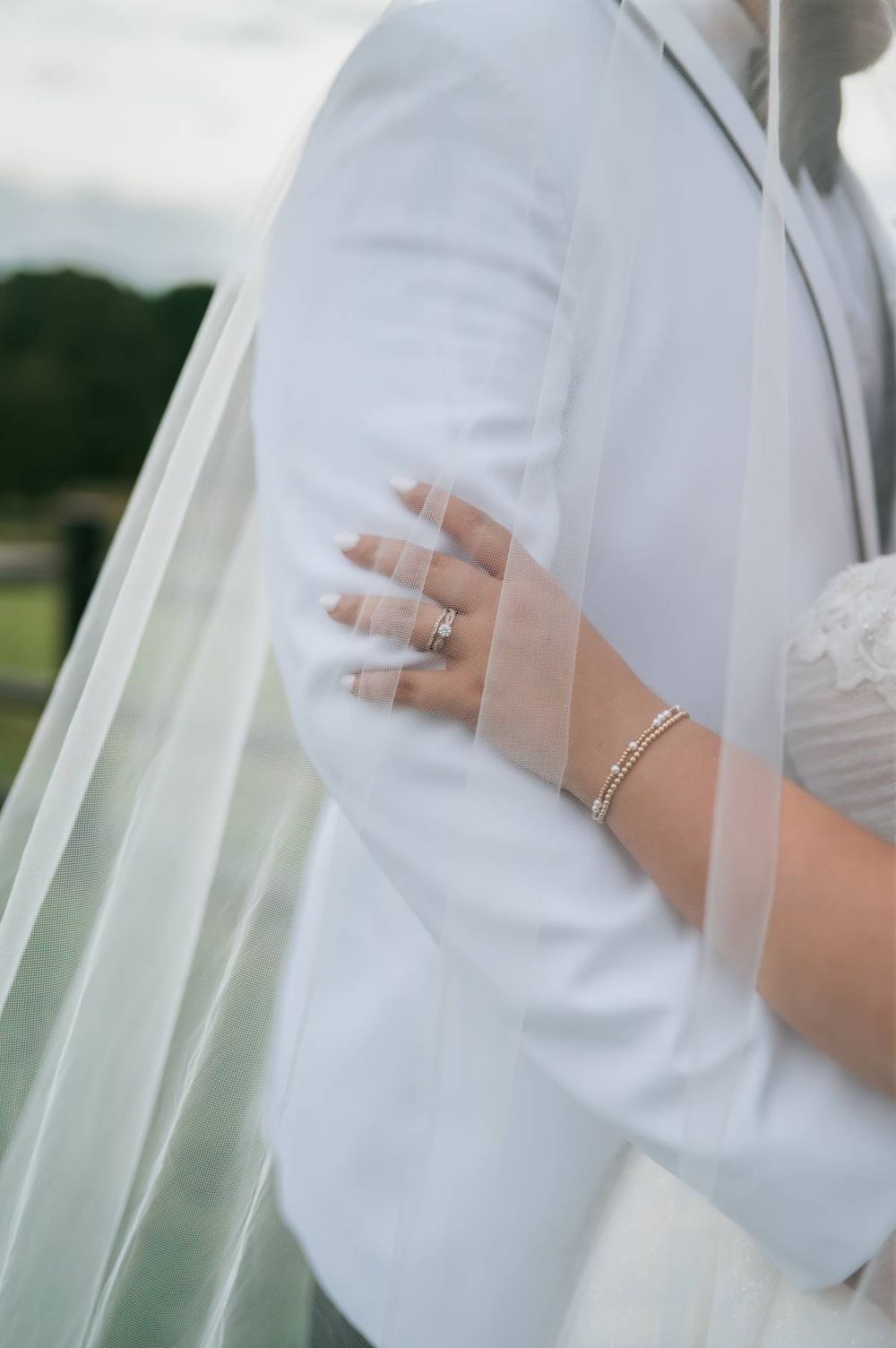 Bride resting hand on groom's white suit jacket, wearing wedding rings and a delicate bracelet, with veil draped softly.