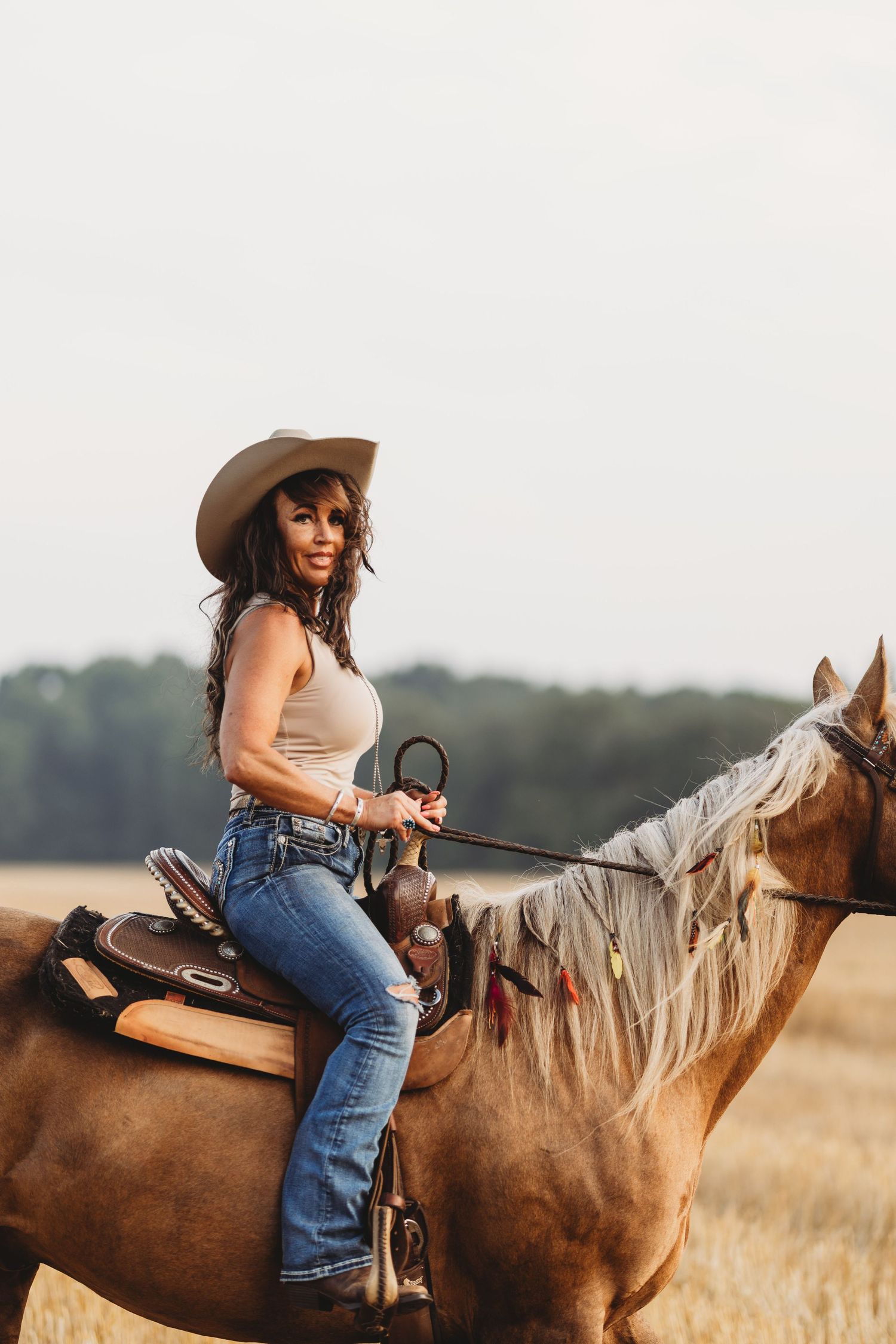 A woman in a cowboy hat and jeans rides a palomino horse through a golden field.