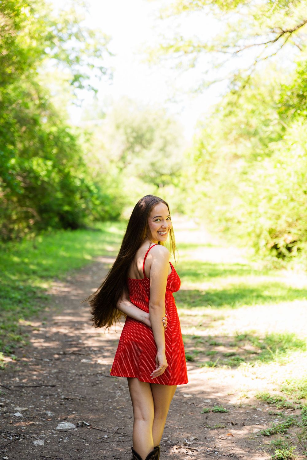 Young woman in red dress walking on a sunlit forest path, smiling over her shoulder with long flowing hair.