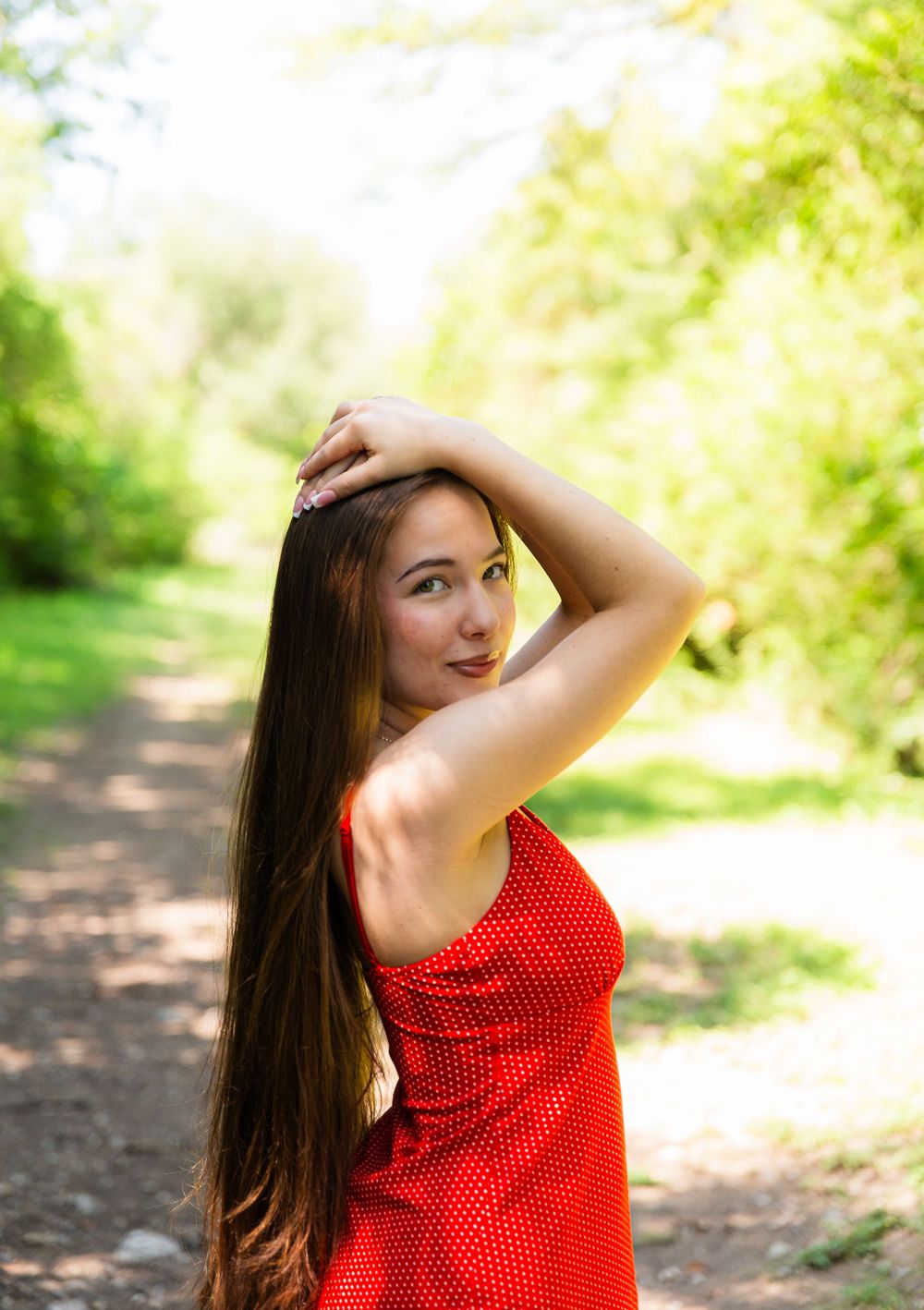 Young woman in red polka dot dress posing outdoors on a sunny forest path with long brown hair.