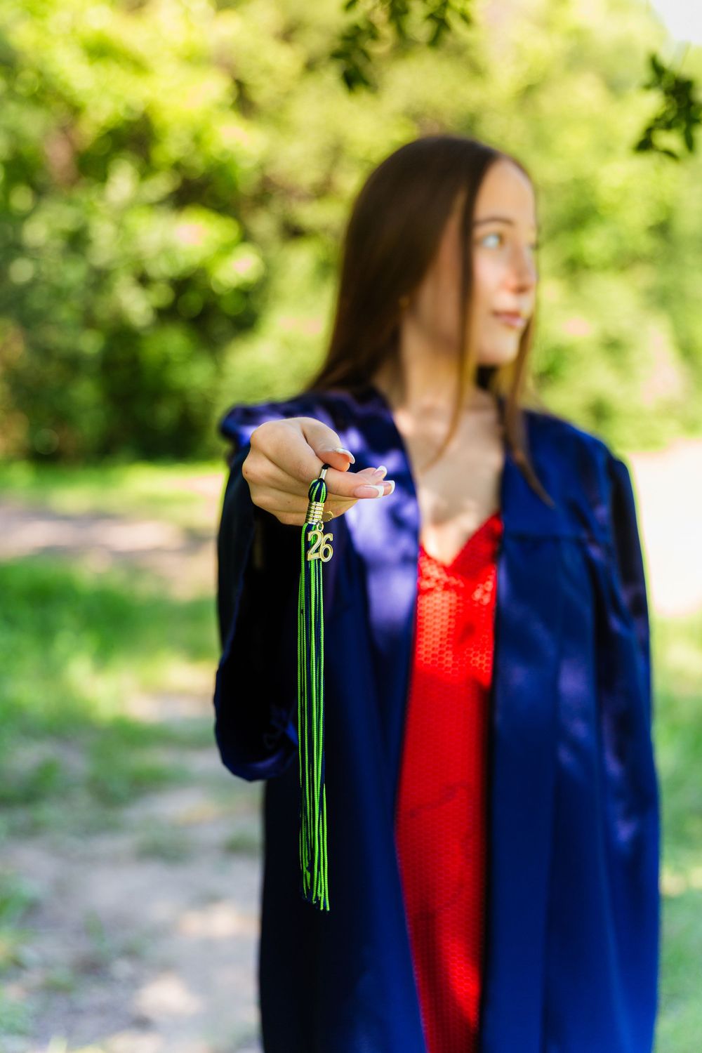 A female graduate in blue gown and red dress holds out a green graduation tassel with charm toward the camera outdoors.