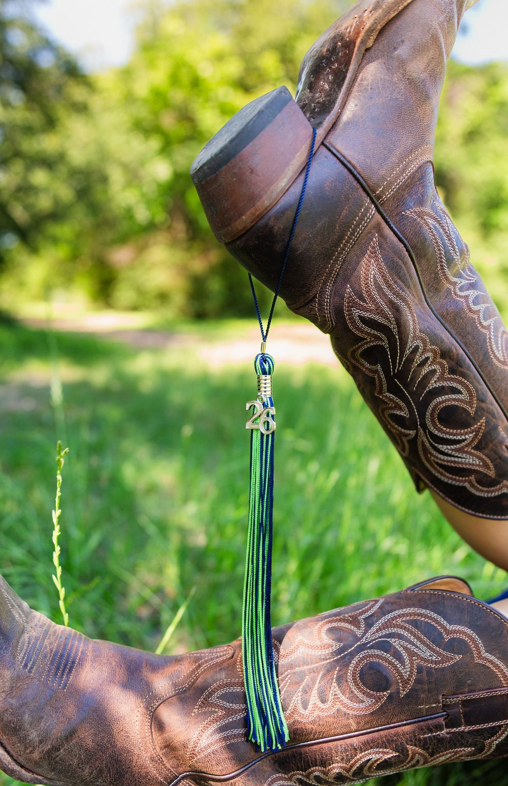 Brown cowboy boots with a green tassel graduation charm hanging between them outdoors.