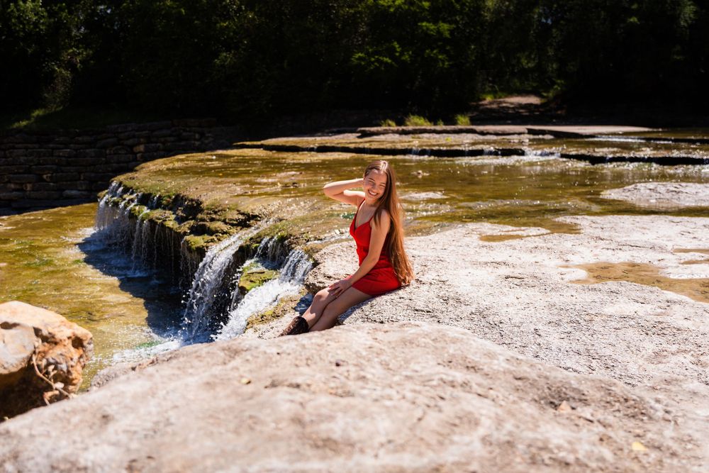 Woman in red swimsuit relaxing on rocky ledge beside a natural waterfall and serene pool surrounded by lush trees.