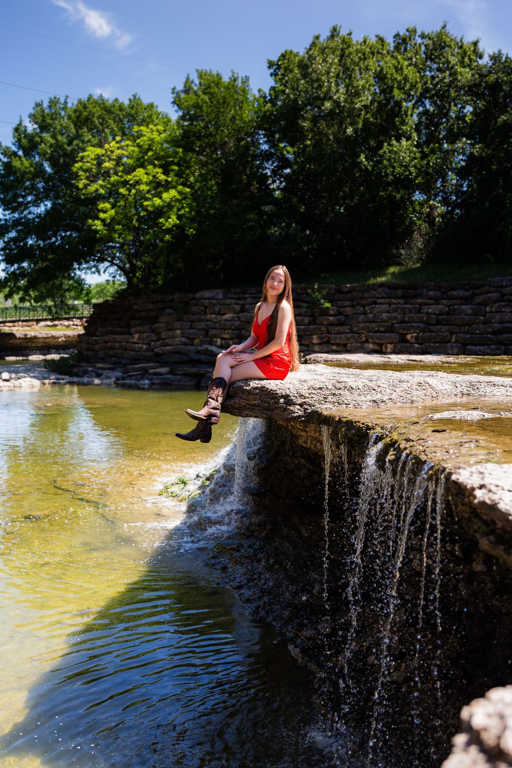 Young woman in red dress sitting on stone ledge over a waterfall in a scenic outdoor park setting.