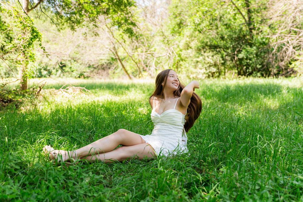 Young woman in white dress sits relaxing on green grass in a sunny park.