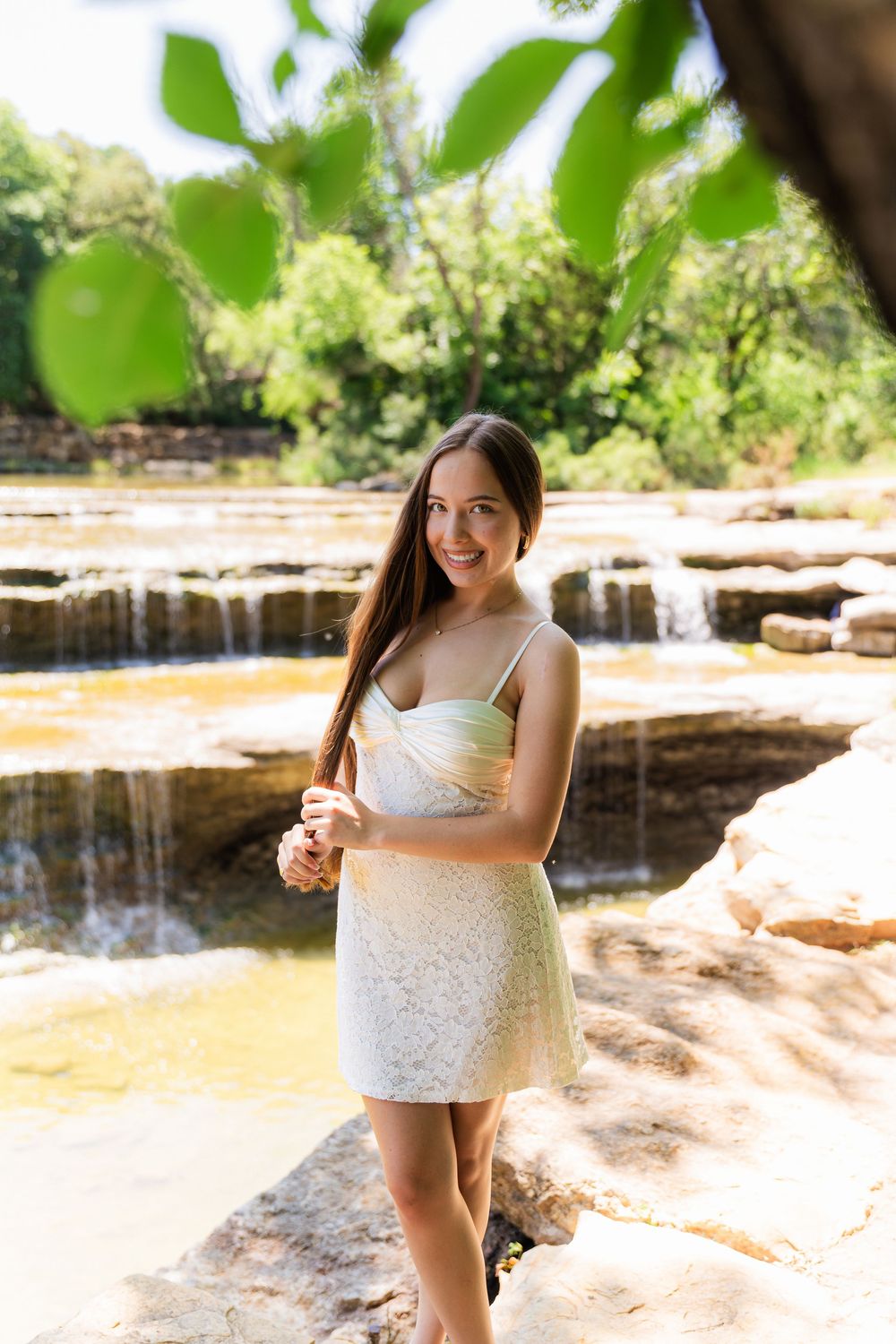 Young woman in white lace dress smiling by a rocky waterfall surrounded by lush green trees.