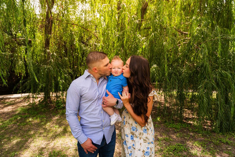 A couple kisses their baby boy under a weeping willow tree in a bright outdoor family portrait.