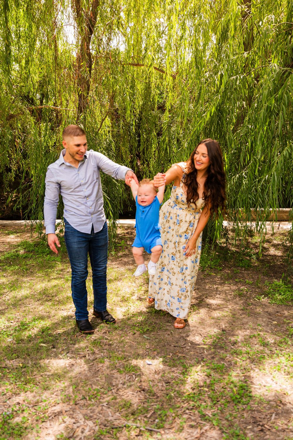 Happy young family with toddler son playing outdoors under a weeping willow tree on a sunny day.