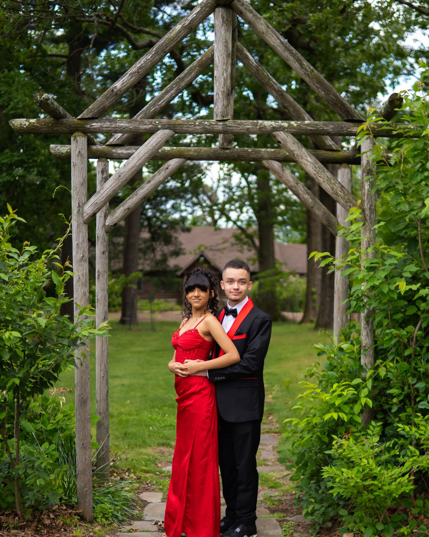 Couple in formal wear posing under rustic wooden arbor surrounded by lush greenery, woman in red gown, man in black tuxedo.