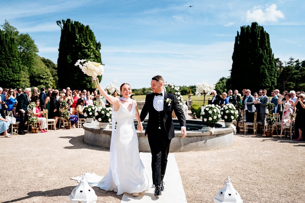 Long panoramic shot captures elegant outdoor garden wedding ceremony with guests seated on white chairs and blooming flowers.