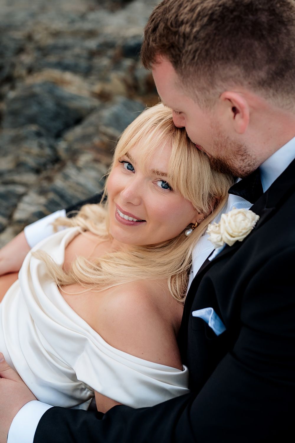 A romantic wedding photoshoot shows the couple in white and black formal attire against a rocky backdrop in natural lighting.