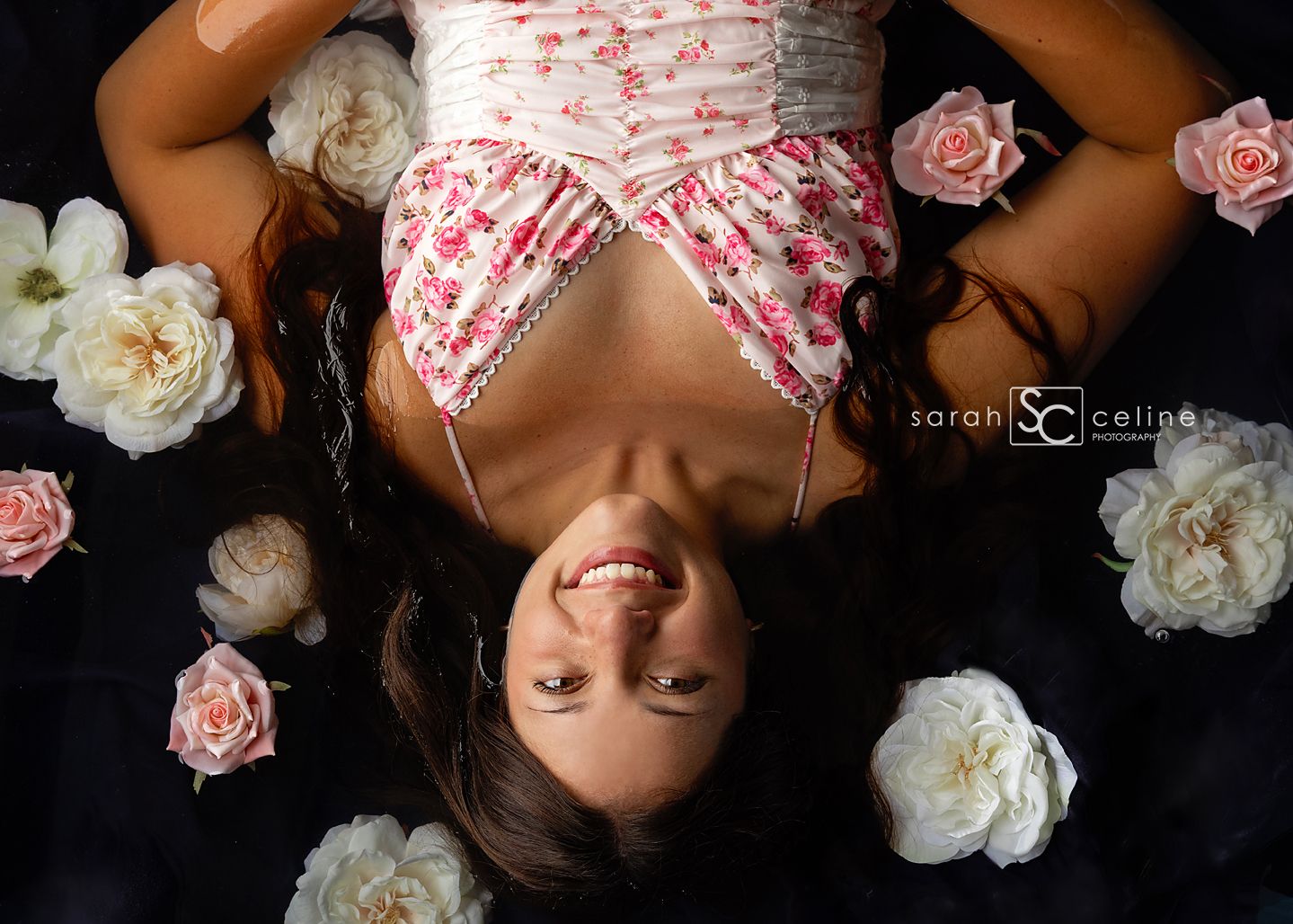 Young woman in floral dress lying among pink and white roses, smiling up at camera, aerial view photography.