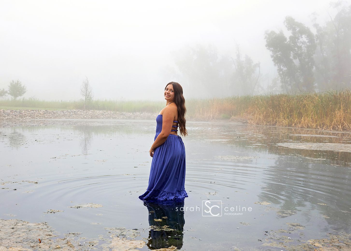 Woman in blue gown standing in misty lake water during outdoor portrait photography session.